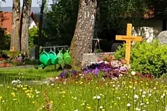 Blick auf einen Friedhof. Ein Holzkreuz, vier grüne Giesskannen und eine blühende wiese und Bäume